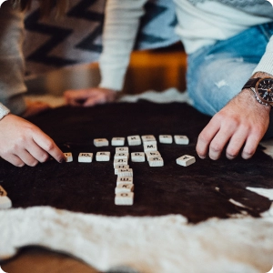 Twee mensen spelen een woordspel op een tapijt met patronen en leggen houten tegels neer om woorden te vormen. De scène is gezellig en ontspannen, met casual kleding.