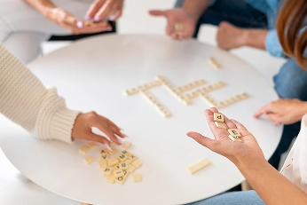 Vier mensen zitten rond een tafel en spelen een woordspel met tegels. Ze leggen tegels neer en spellen ‘THE’ in het midden. De scène voelt informeel en boeiend aan.